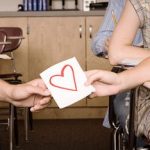 Symbolische Darstellung der ersten Liebe: männliche und weibliche Hand halten gemeinsam an Papier mit rotem Herz in Klassenzimmer