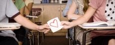 Symbolische Darstellung der ersten Liebe: männliche und weibliche Hand halten gemeinsam an Papier mit rotem Herz in Klassenzimmer