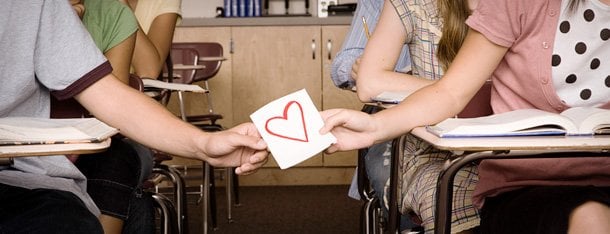 Symbolische Darstellung der ersten Liebe: männliche und weibliche Hand halten gemeinsam an Papier mit rotem Herz in Klassenzimmer