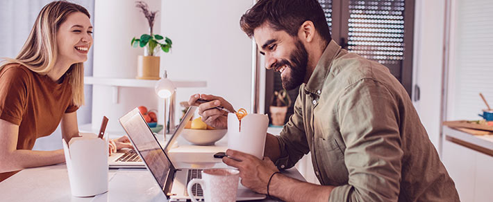 Ein Paar sitzt gemeinsam in der eigenen Wohnung im Homeoffice, für ihre Beziehung hat es sich positiv ausgewirkt, denn sie lachen