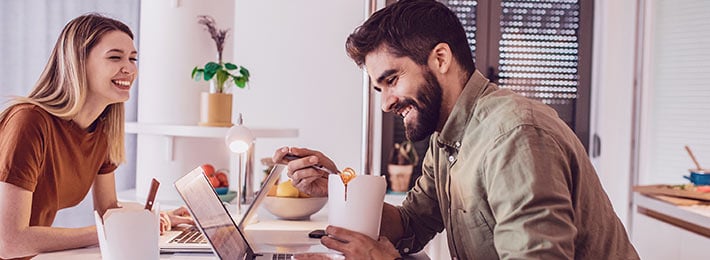 Ein Paar sitzt gemeinsam in der eigenen Wohnung im Homeoffice, für ihre Beziehung hat es sich positiv ausgewirkt, denn sie lachen