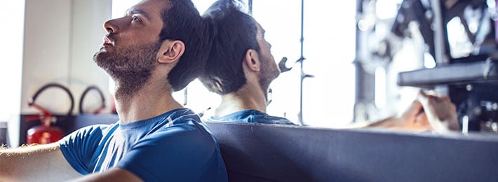 Ein Mann sitzt erschöpft im Fitnessstudio, er zweifelt an seiner eigenen Attraktivität und hat beim Dating oft Body Anxiety