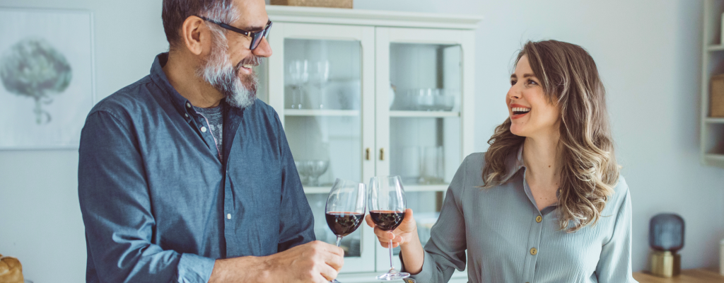 Ein Mann mit grau meliertem Bart und Brille und eine Frau mit schulterlangen, brünetten Haaren kochen gemeinsam in einer weißen Küche und stoßen mit einem Glas Rotwein an.